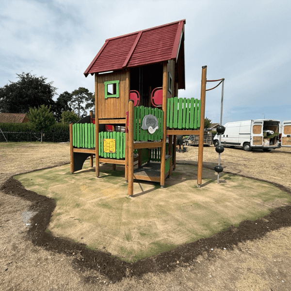 Children’s wooden play tower with slide and activity panels, installed on new playground surfacing in a partially developed outdoor area .