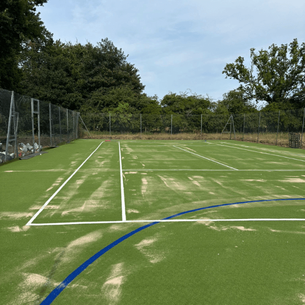 A sports court with bright green artificial turf, white court markings, and scattered patches of loose sand across the surface.